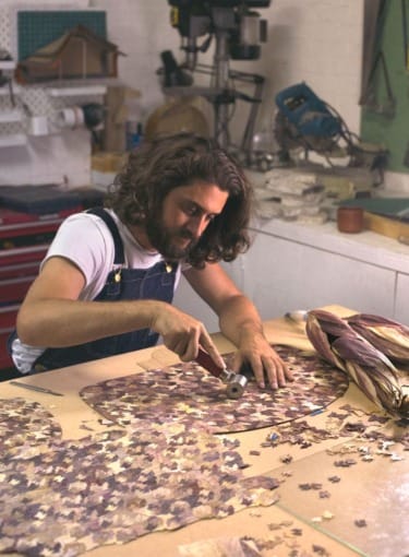 Man crafting with dried leaves at workbench in workshop, using hammer and punch on natural material.