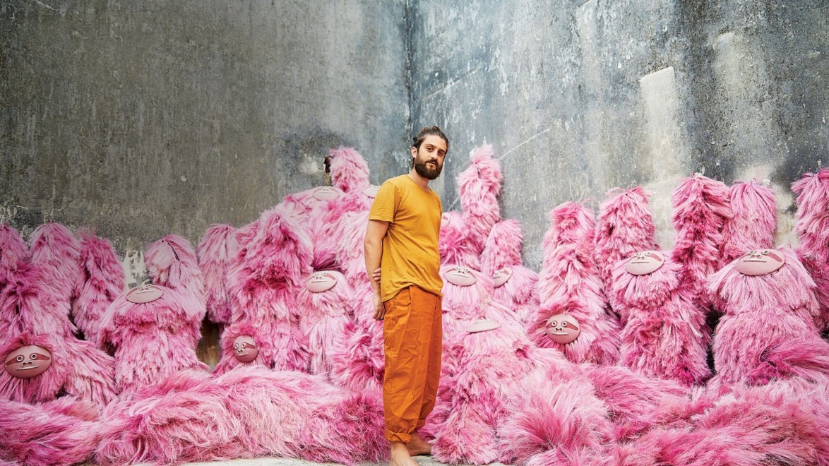 Man in orange outfit standing among large pink furry creatures in an industrial room.