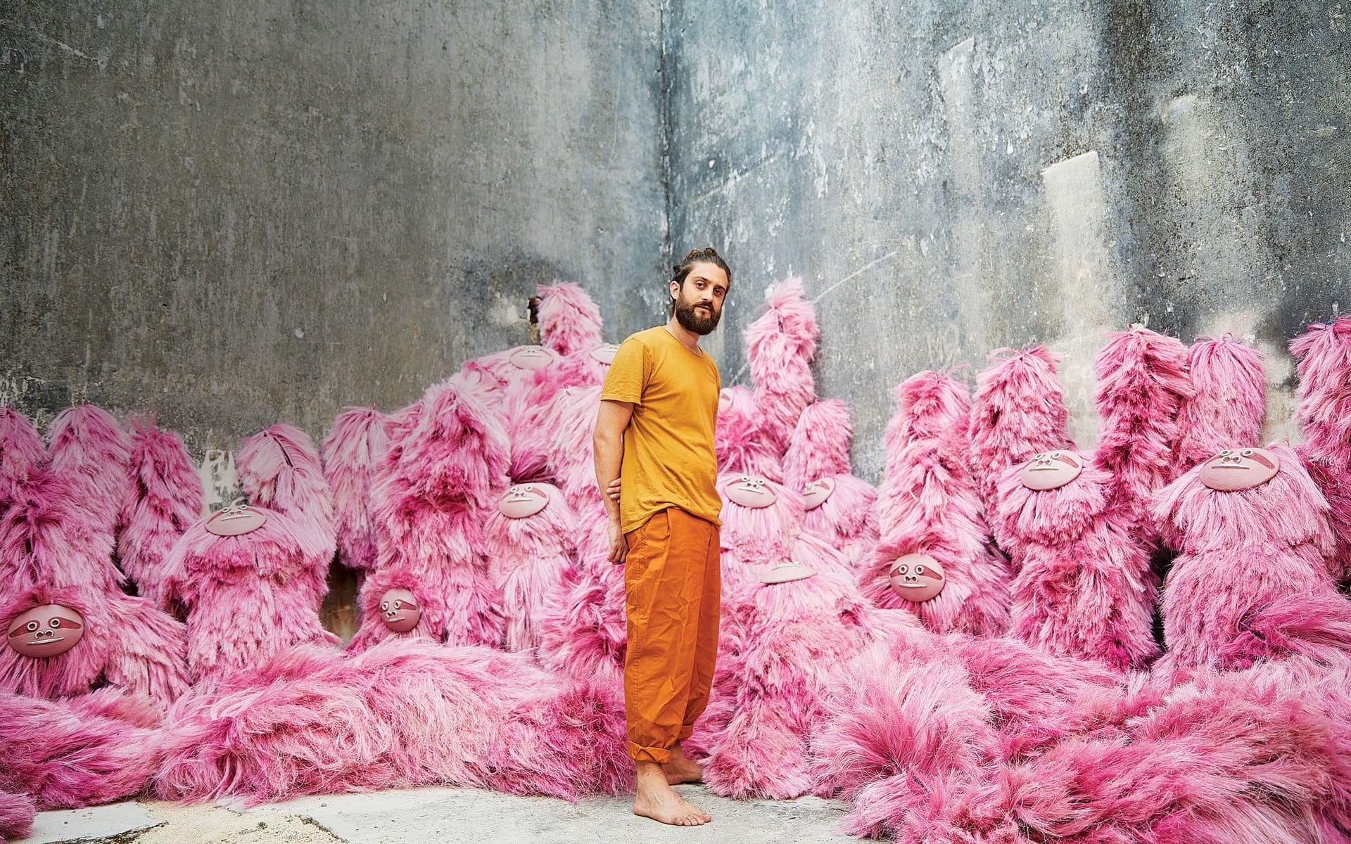 Man in orange outfit standing among large pink furry creatures in an industrial room.