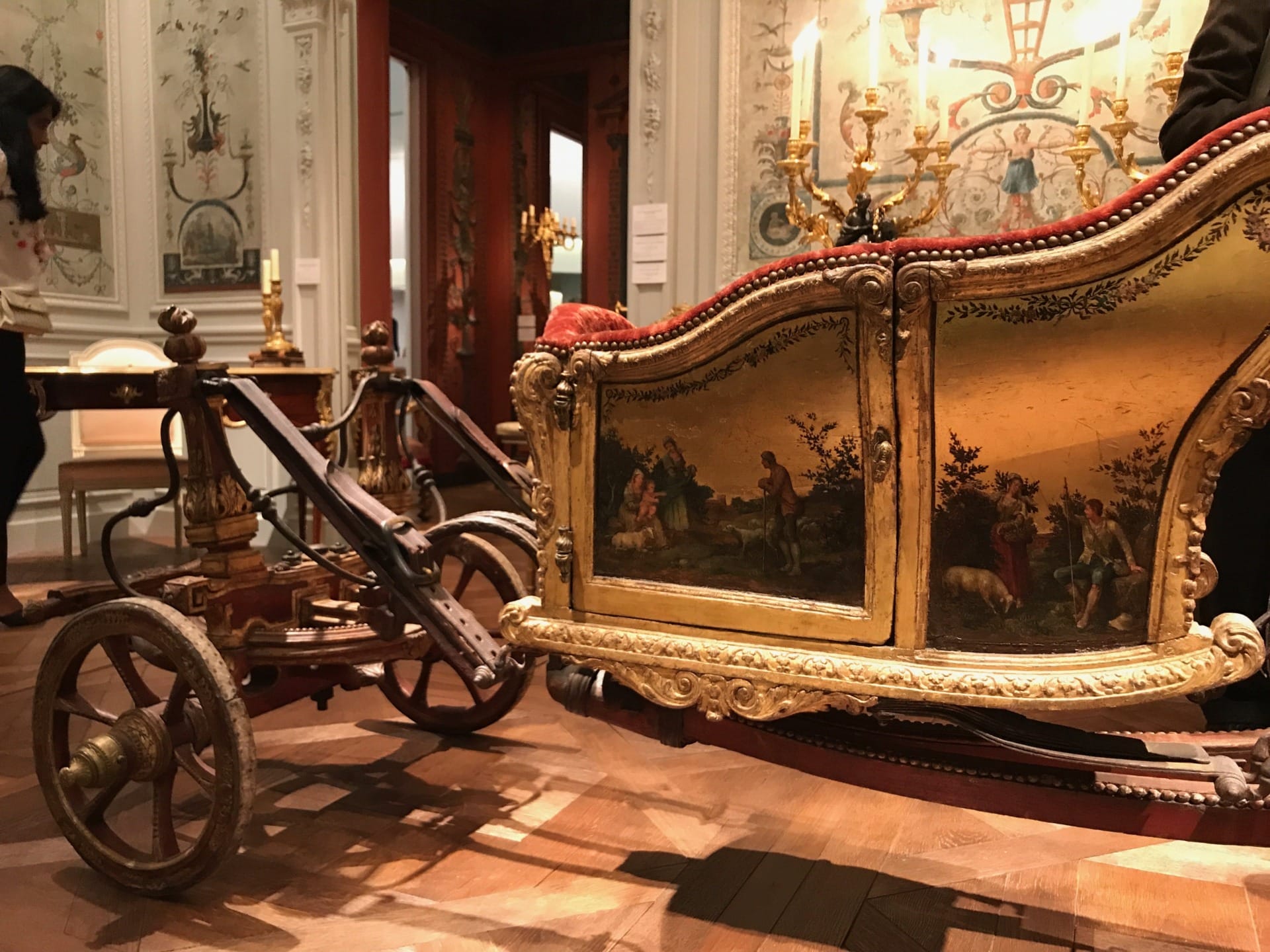Antique ornate carriage with intricate paintings displayed in a museum setting with wooden flooring and decorative walls.