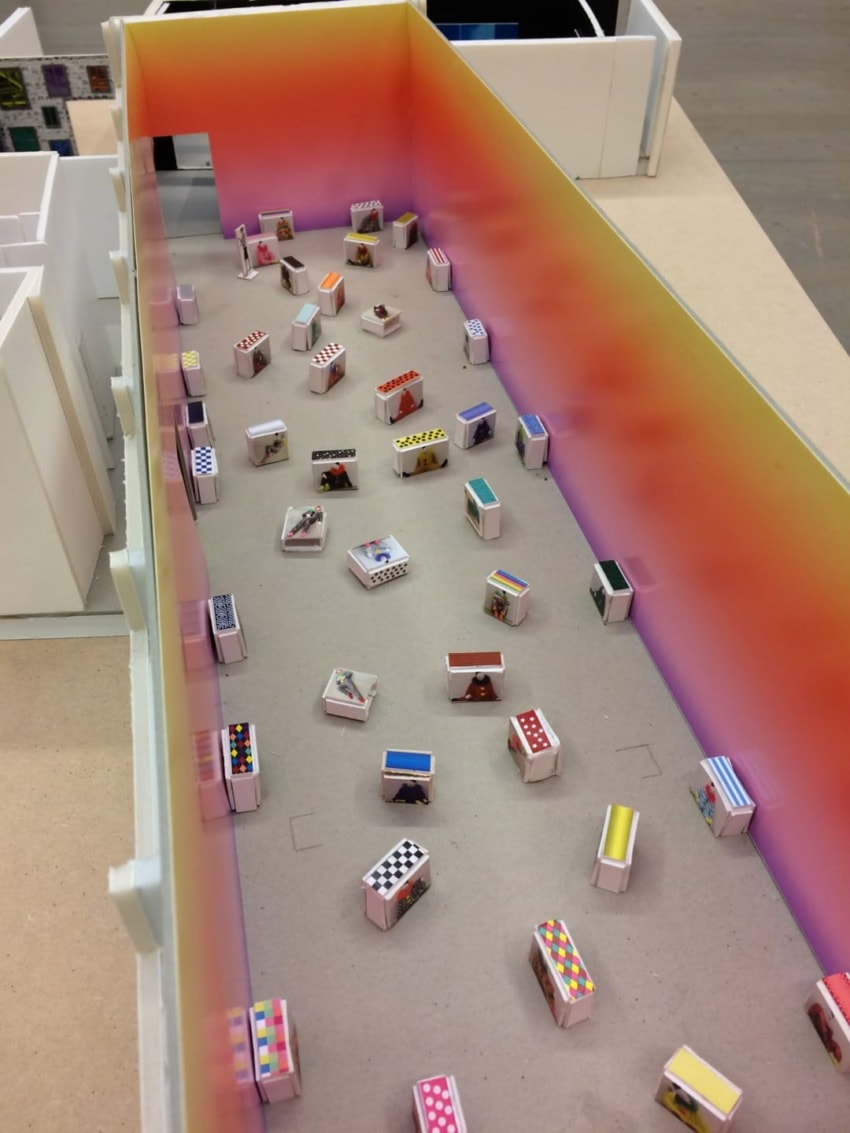 Model of an art gallery with miniature decorated boxes arranged on the floor, surrounded by colorful gradient walls.