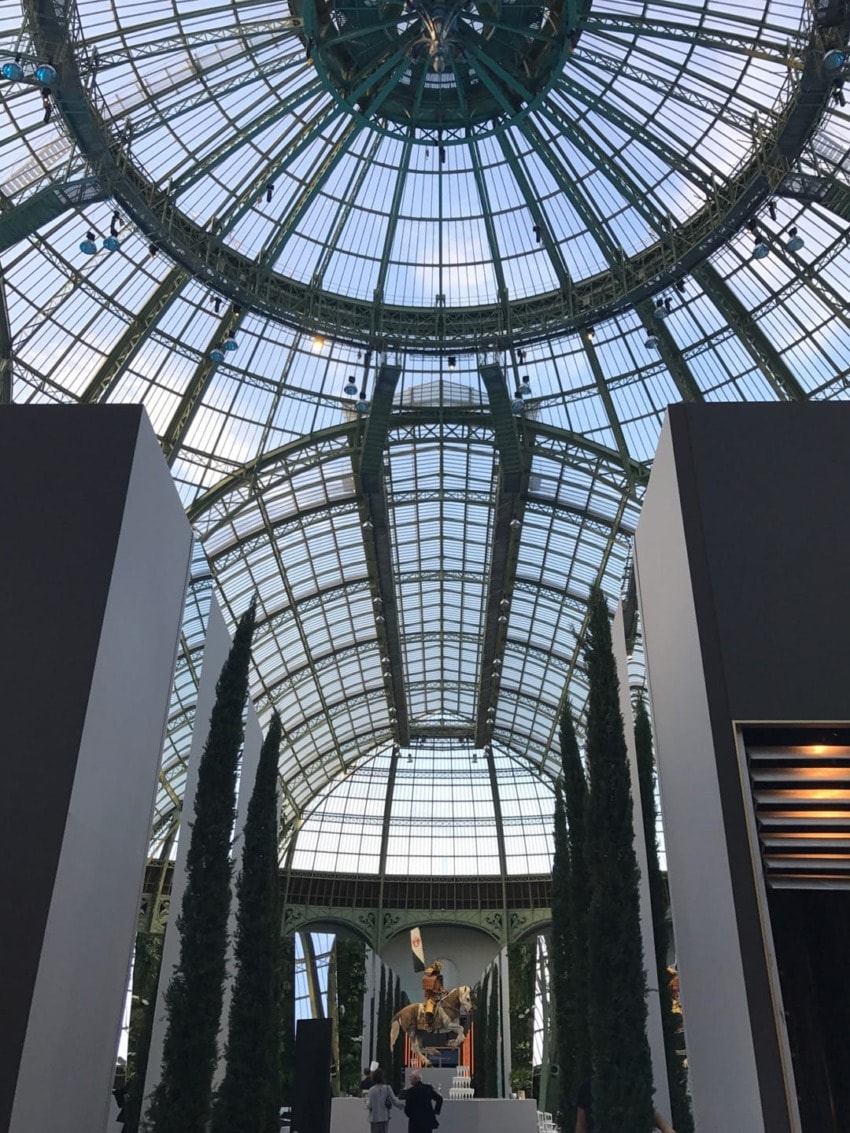 Interior view of a grand glass ceiling and metallic structure with tall trees lining an elegant space at an exhibition.