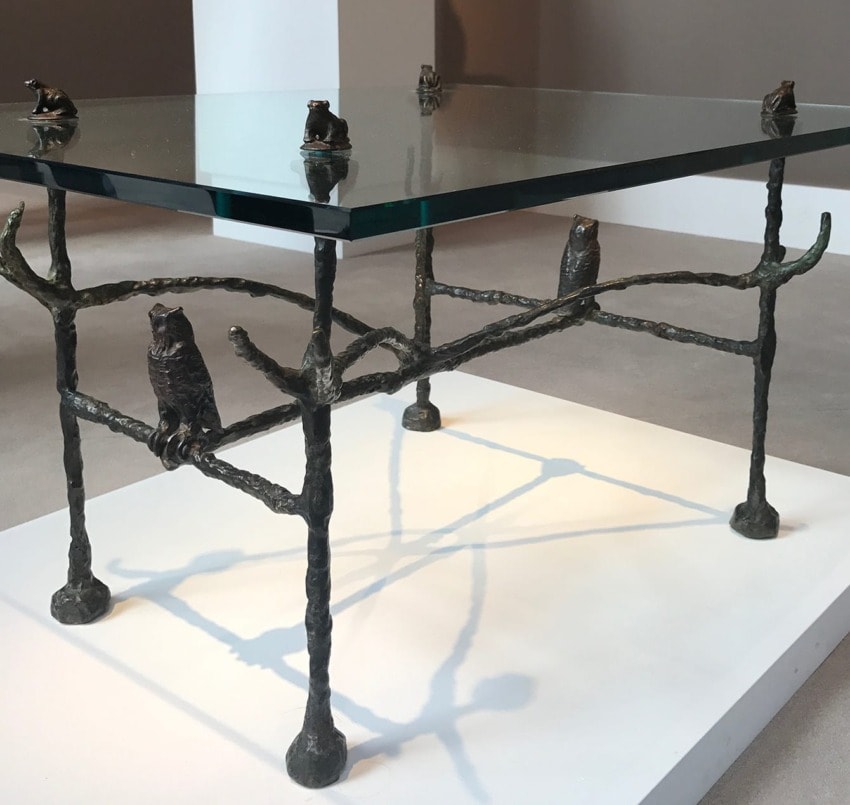 Glass table with sculpted animal figures on metal legs displayed on a white platform in a gallery setting.