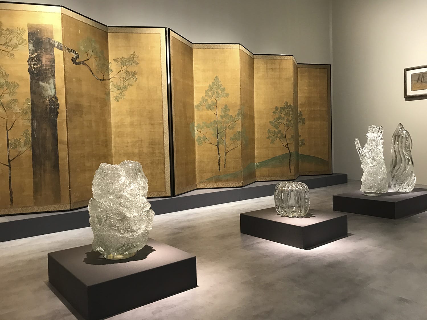 Museum exhibit featuring glass sculptures displayed in front of a gold-painted folding screen with tree designs.
