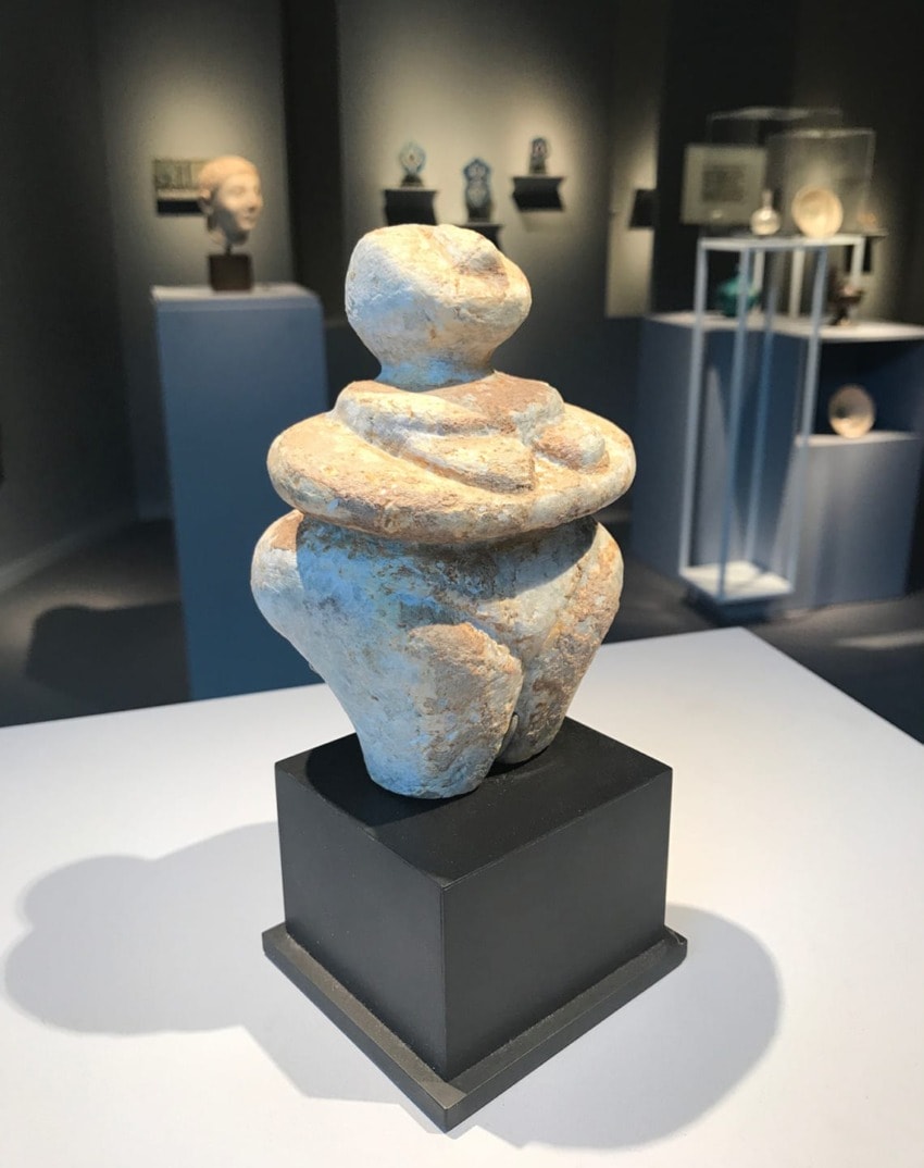 Abstract stone sculpture on display in an art gallery setting with several other artifacts in the background.