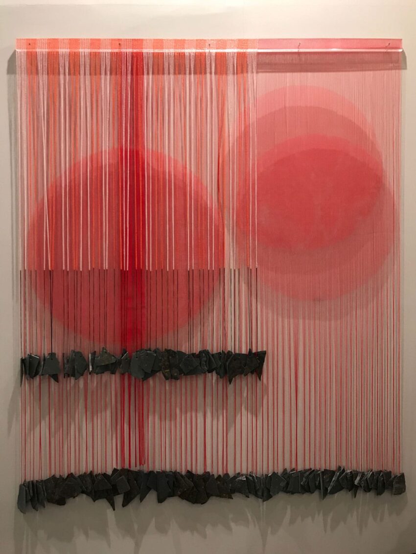 Abstract art piece with vertical red threads, two circular shapes, and black stones aligned at the bottom.