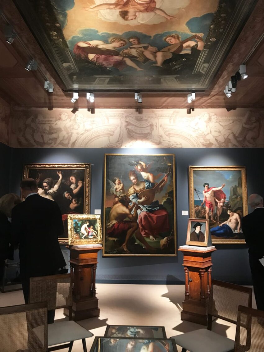 Art gallery interior with classical paintings and ornate ceiling, several people viewing the artworks in a well-lit room.