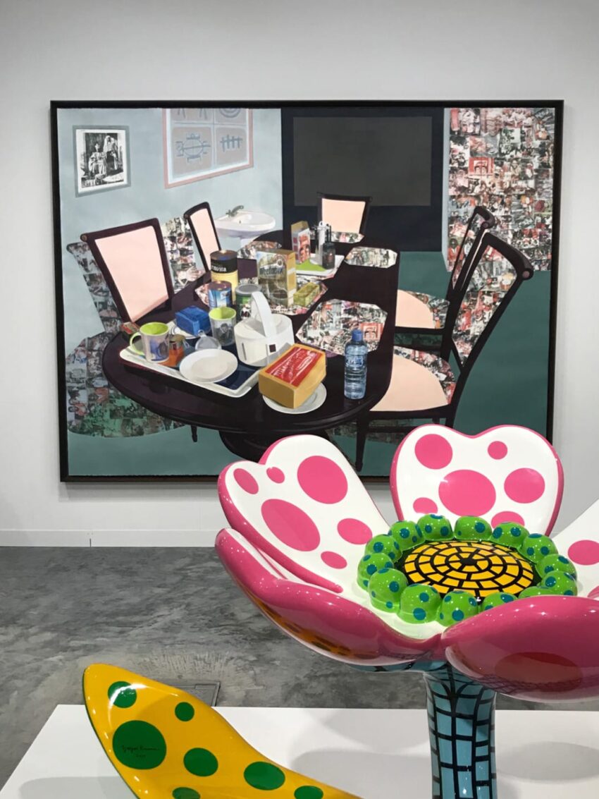 Colorful abstract flower sculpture in foreground with a painting of a dining table with assorted items in the background.
