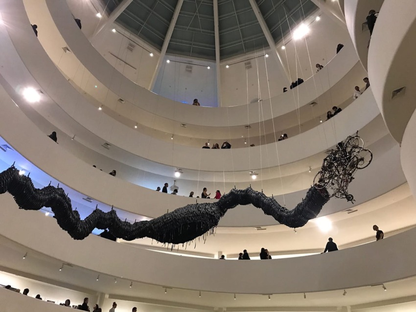 Contemporary art installation inside a multi-level modern museum with visitors observing from the spiraling walkways.