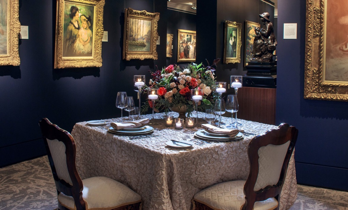 Elegant dining table set with flowers and candles in an art gallery surrounded by framed paintings on dark walls.