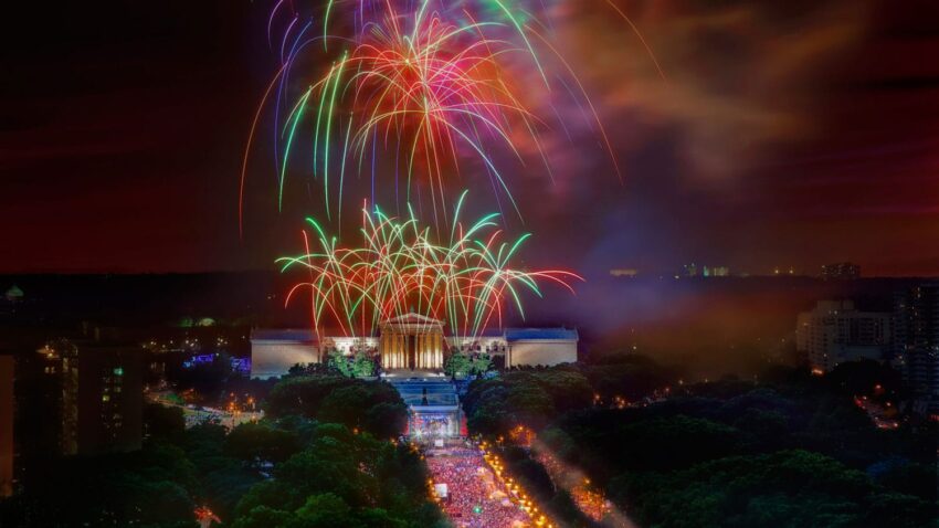 Colorful fireworks light up the night sky over a large building surrounded by trees and a bustling crowd below.
