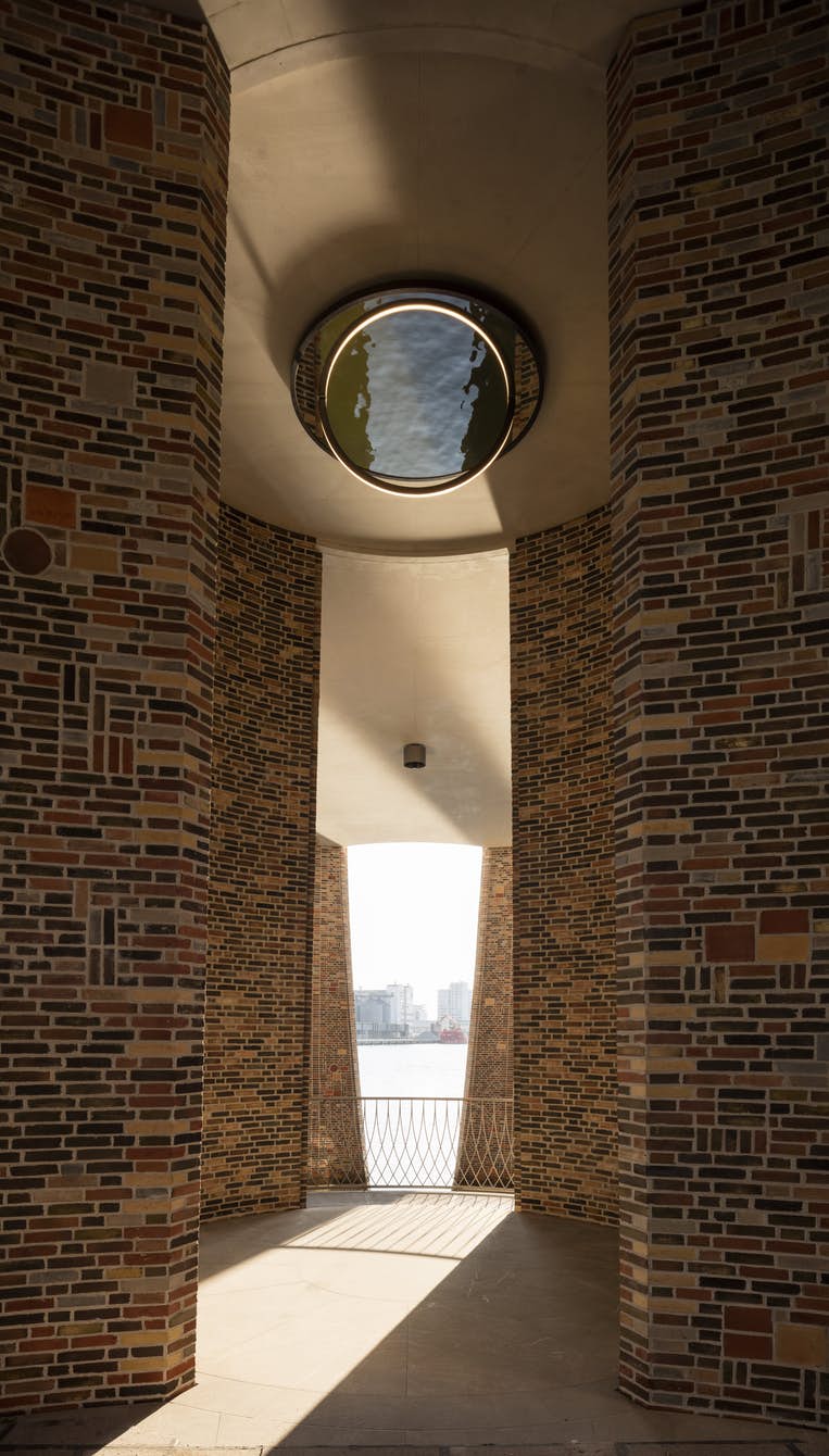 Brick walls encircle a circular skylight, illuminating a passageway with a view of a river and distant cityscape.