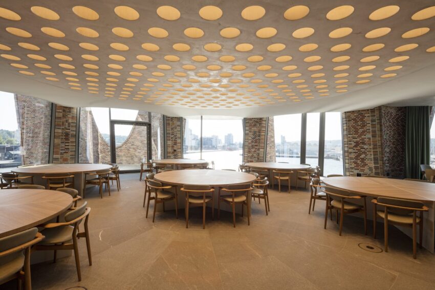 Modern circular room with large round tables, brick walls, and patterned ceiling overlooking a cityscape through large windows.