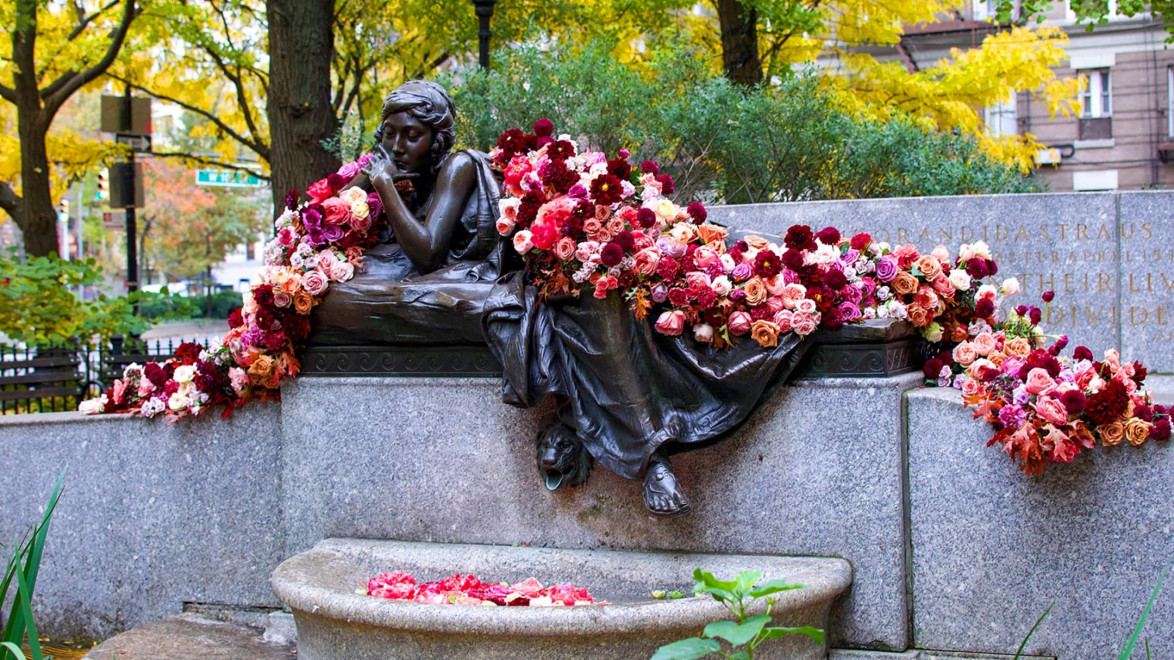 Statue in a park adorned with vibrant flowers, surrounded by autumn trees and a city street in the background