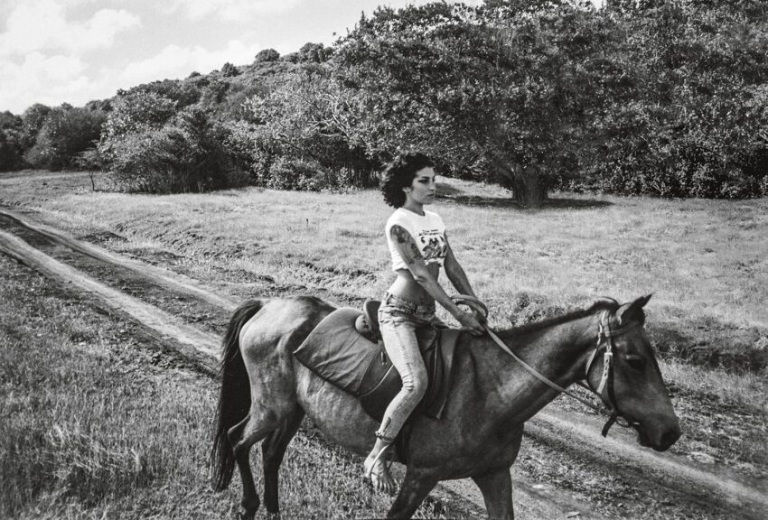 A person riding a horse on a dirt path in a grassy landscape with trees in the background, captured in black and white.