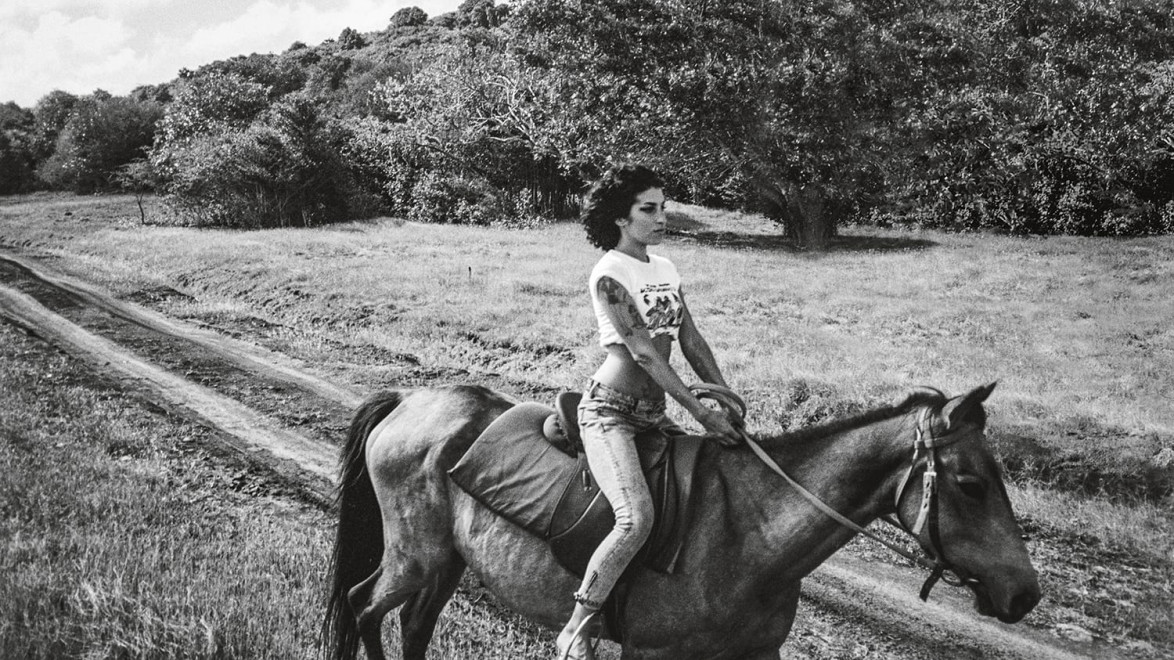 A person riding a horse on a dirt path in a grassy landscape with trees in the background, captured in black and white.