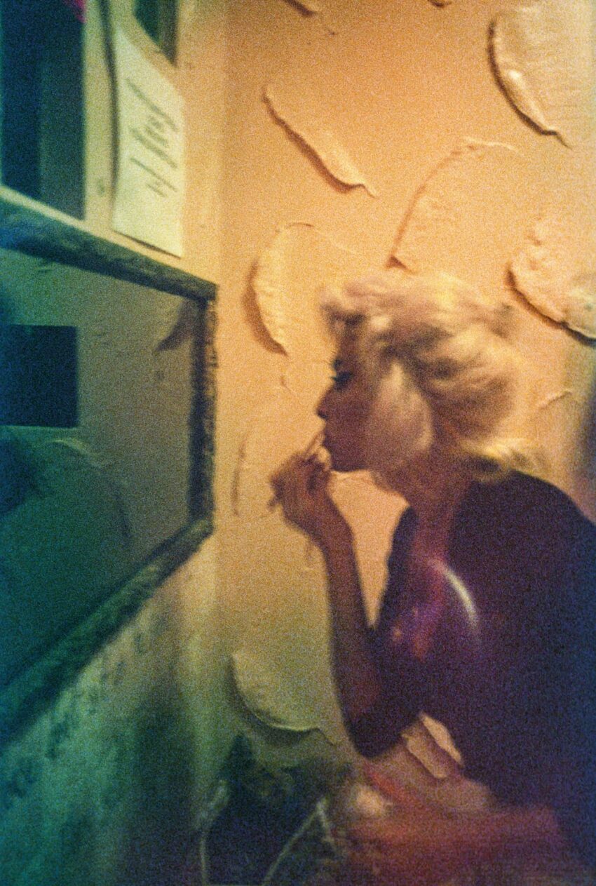 Person applying makeup in a dimly lit room, standing in front of a mirror with textured walls in the background.