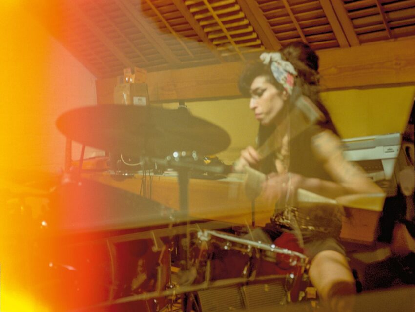 Woman passionately playing drums in a dimly lit room with a noticeable light flare effect.