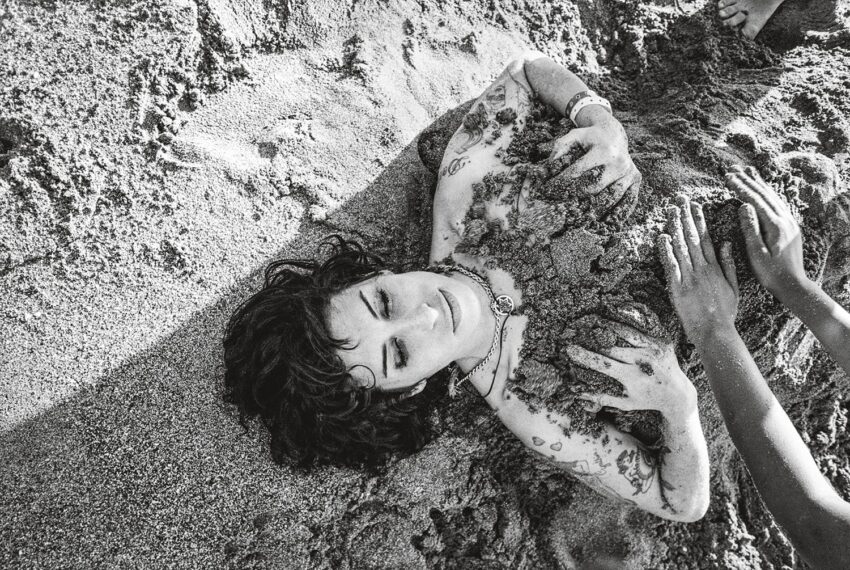 Person lying on sand at beach, partially covered in sand, with tattoos on arms and chest, surrounded by hands.