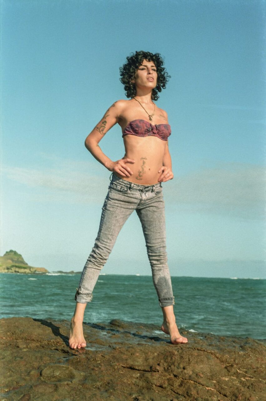 Person with curly hair standing on rocks by the sea, wearing a bikini top and jeans, looking into the distance.