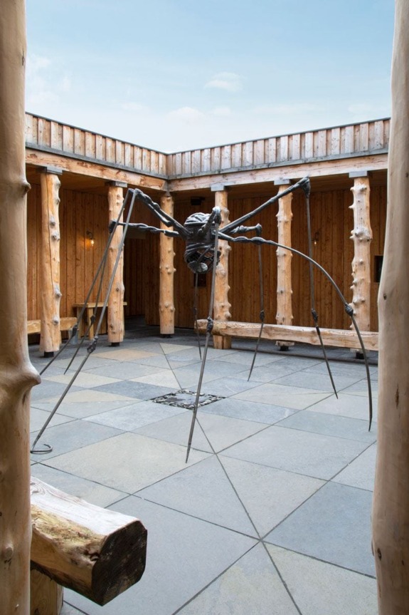 Metallic spider sculpture in a wooden courtyard with stone flooring, surrounded by rustic wooden pillars under a clear sky.