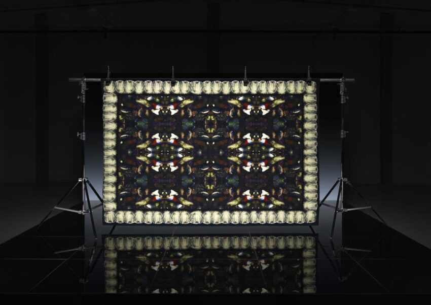 Intricate abstract tapestry with symmetrical patterns, illuminated on a black backdrop, supported by studio stands.