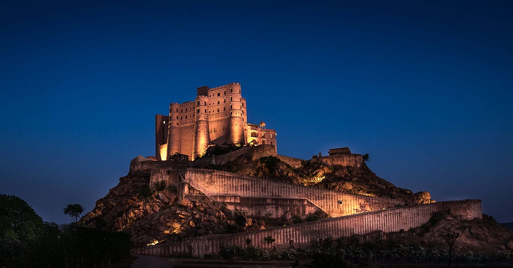 Illuminated fort on a hilltop against a deep blue evening sky, with stone walls and surrounding landscape in view.