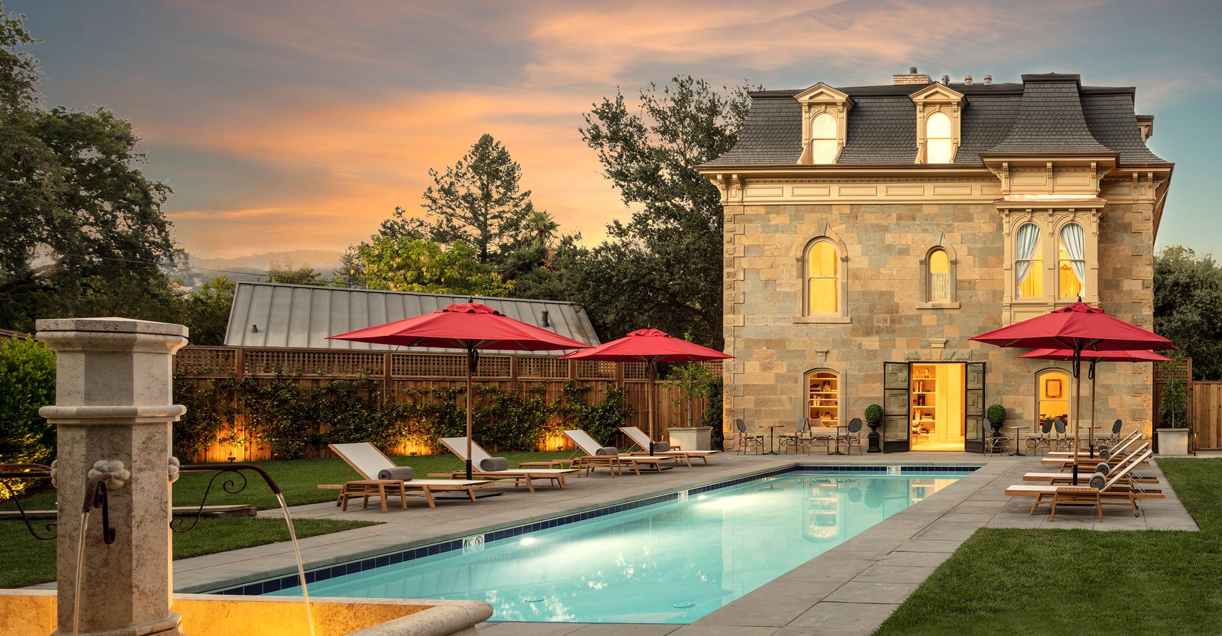 Historic stone house with mansard roof and a long rectangular pool, surrounded by red umbrellas and lounge chairs at sunset.