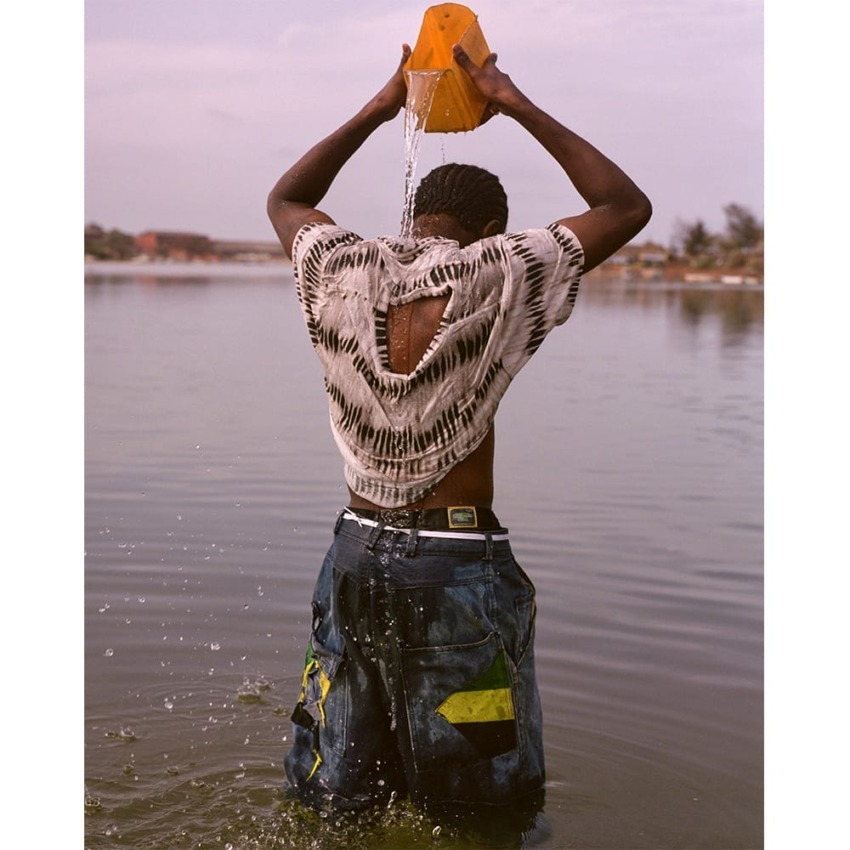 Person standing waist-deep in water, pouring water over their head from a yellow container, wearing a patterned shirt and jeans.