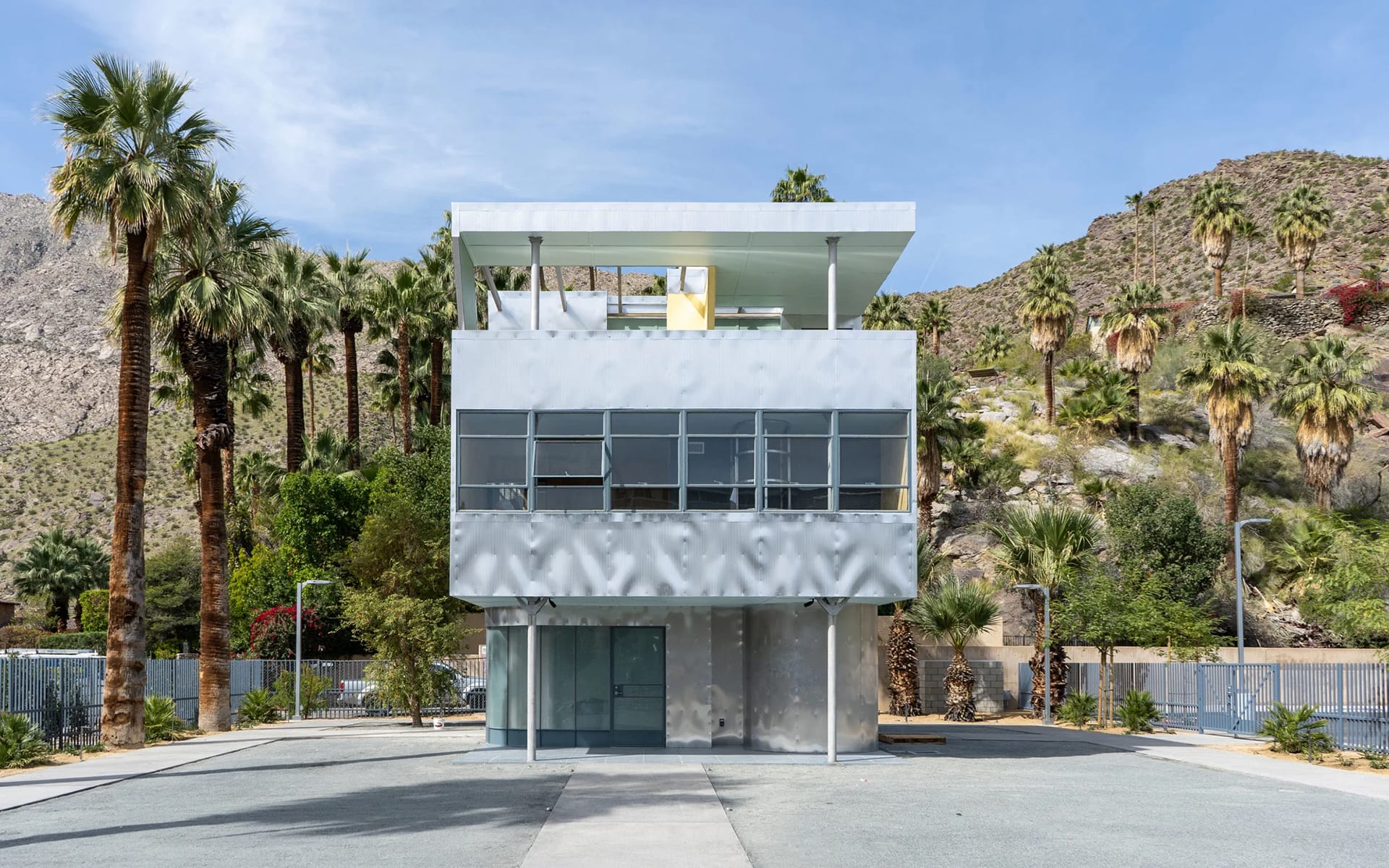 Modern two-story building with glass facade surrounded by palm trees and mountains in a sunny desert landscape.