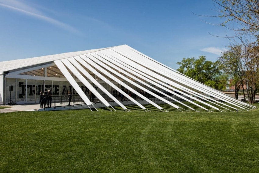 Large open-air white tent with flowing side panels on a lush green lawn under a clear blue sky.