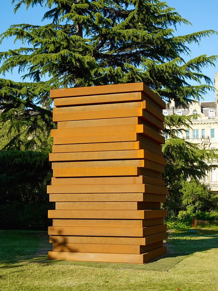 Tall stacked metal sculpture on grass with large tree and buildings in the background under clear blue sky.