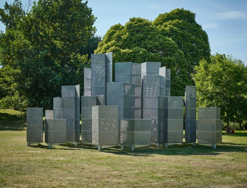 Metallic sculpture composed of geometric panels arranged in a stack, set on a grassy field with trees in the background.