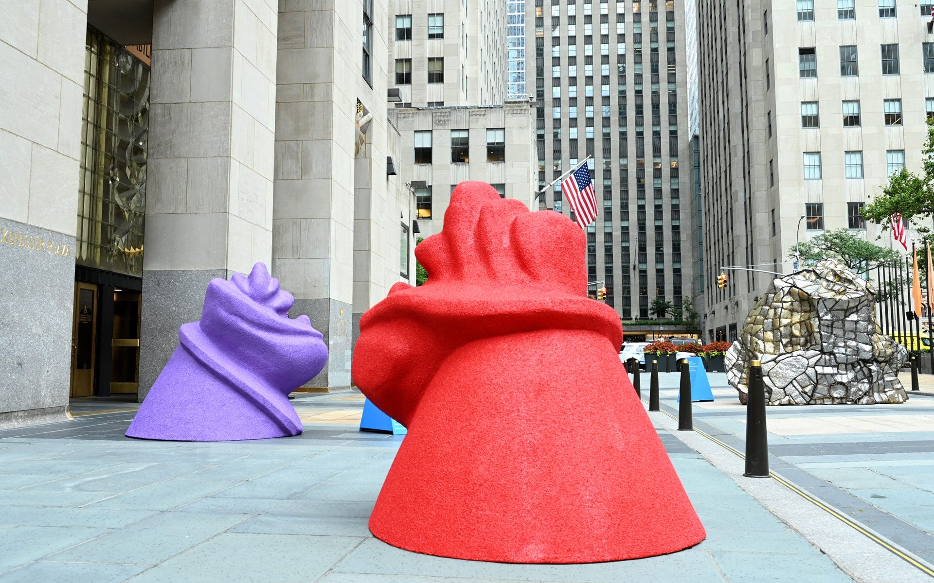 Colorful abstract sculptures displayed in an urban plaza surrounded by tall buildings and American flags in the background.
