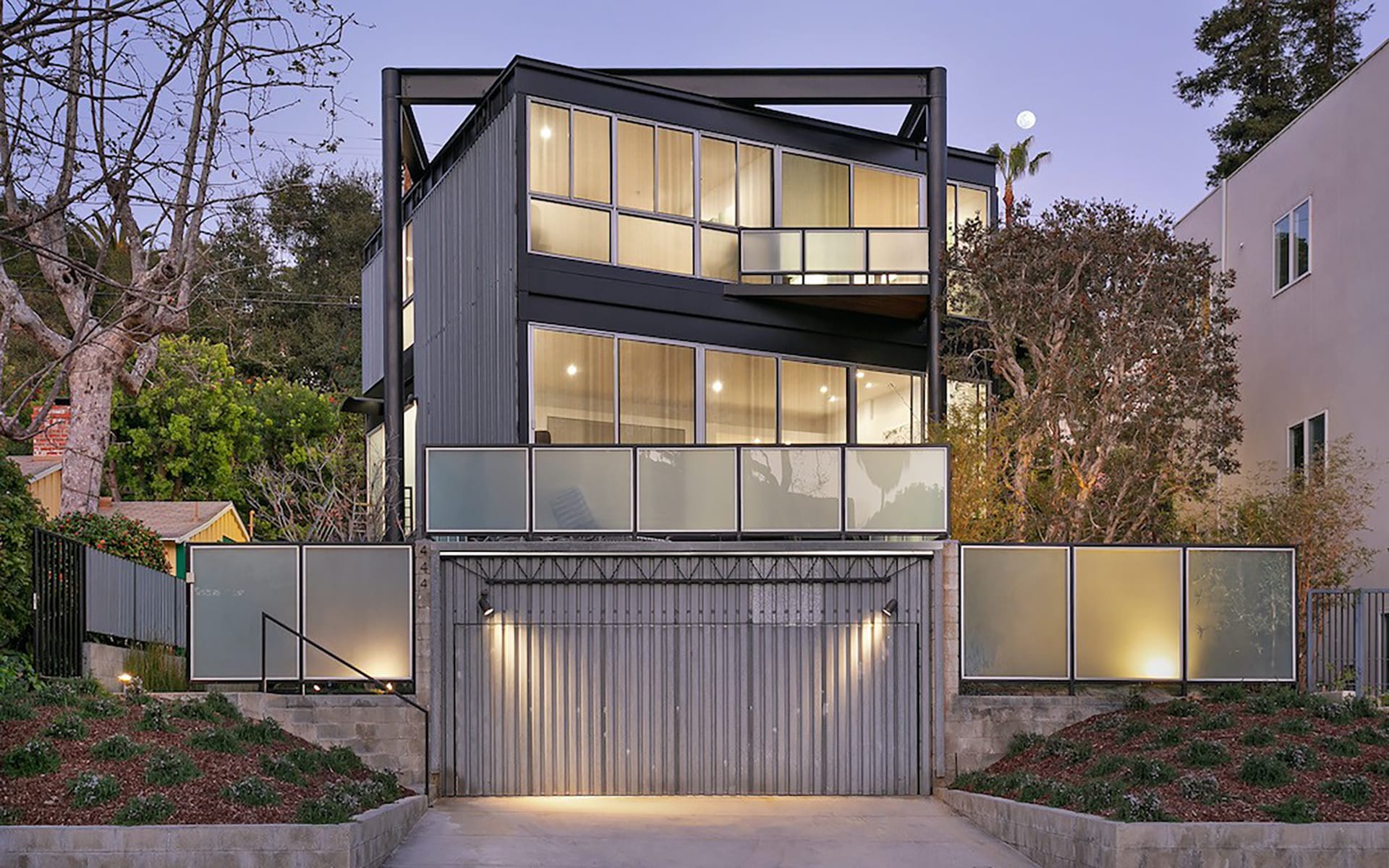 Modern three-story house with large glass windows and a corrugated metal garage door, surrounded by trees and evening lighting.