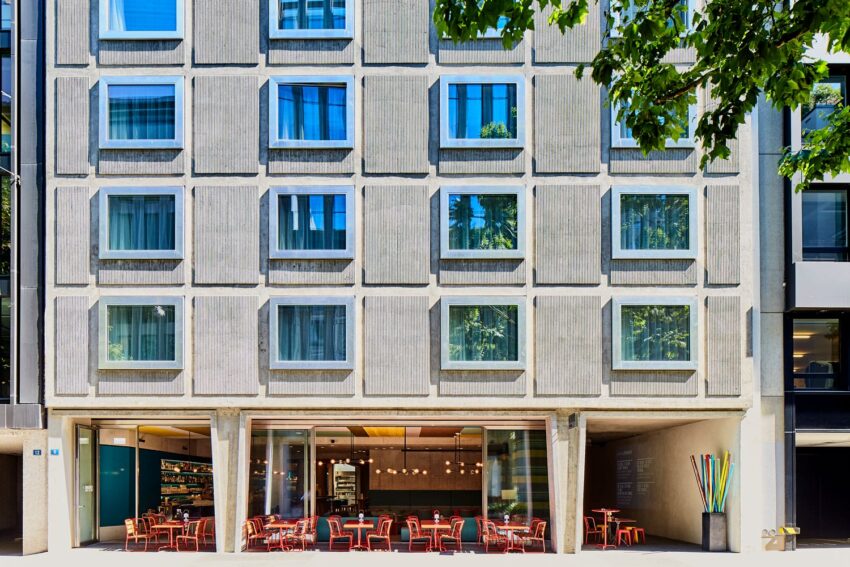 Modern building facade with large windows and outdoor cafe seating under a leafy tree.