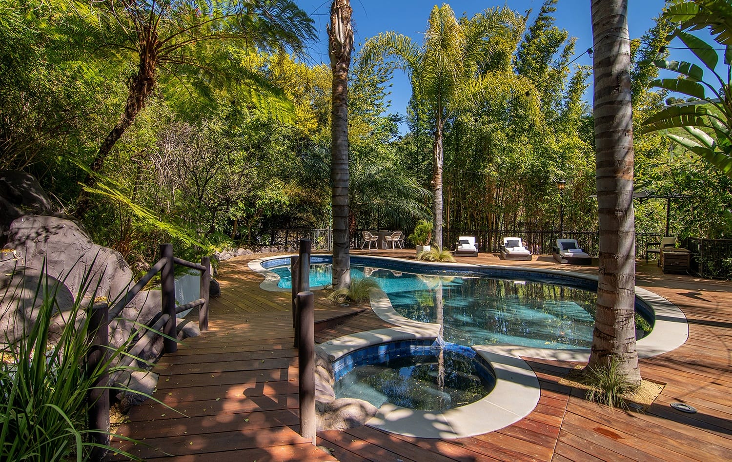 Tropical backyard with a wooden deck, pool, hot tub, palm trees, and lounge chairs on a sunny day.