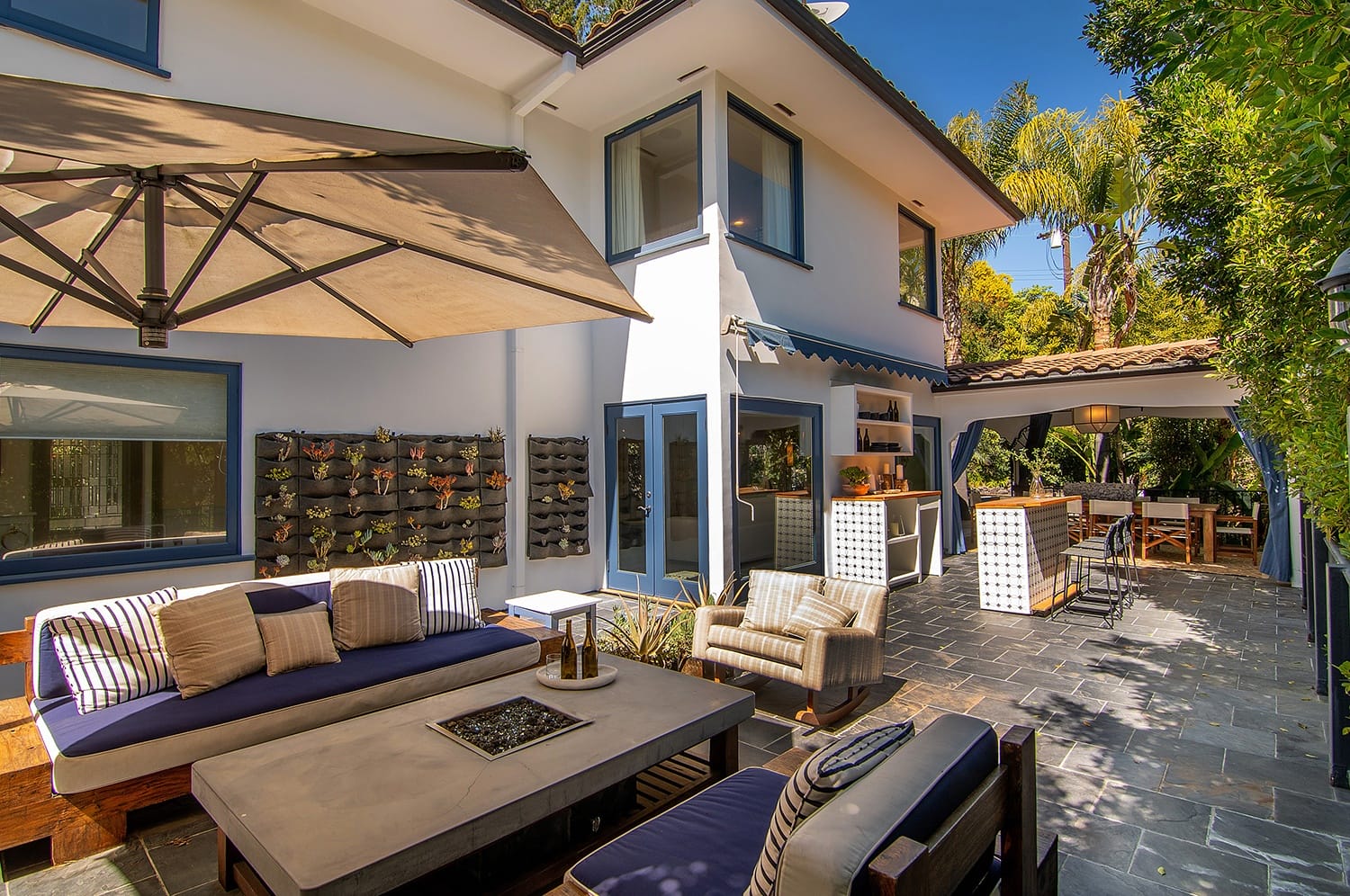 Modern outdoor patio with seating area, umbrella, and outdoor kitchen, surrounded by greenery and a two-story house.