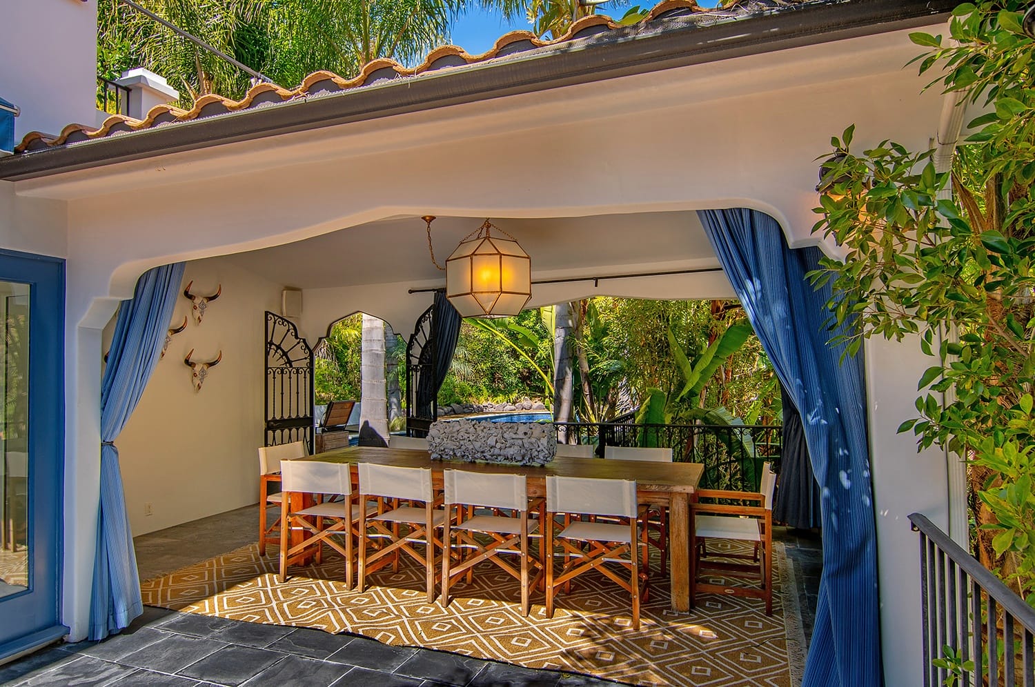 Outdoor dining area with wooden table, white chairs, blue curtains, and decorative lighting surrounded by lush greenery.