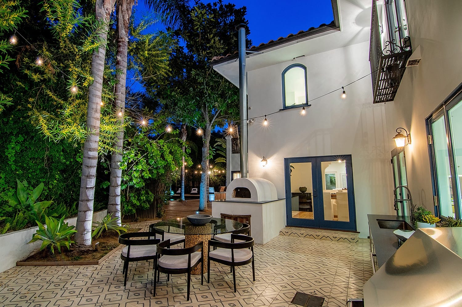 Outdoor patio with string lights, dining table, and wood-fired oven surrounded by lush greenery and a modern white house.