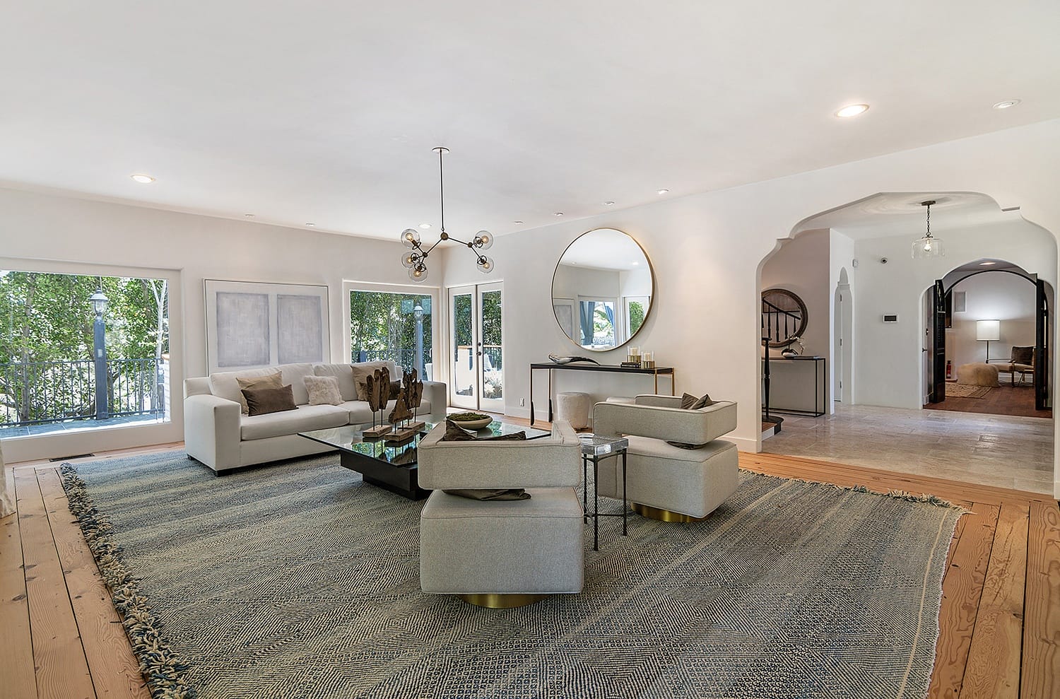 Spacious living room with modern furniture, large rug, round mirror, and ample natural light from large windows.