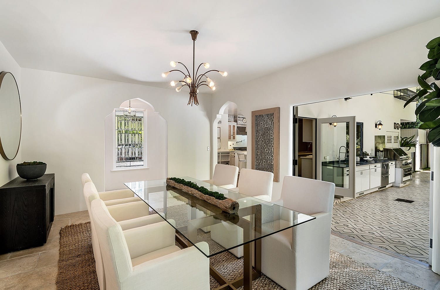 Modern dining room with glass table, white chairs, large chandelier, and view into a stylish kitchen with patterned floor tiles.