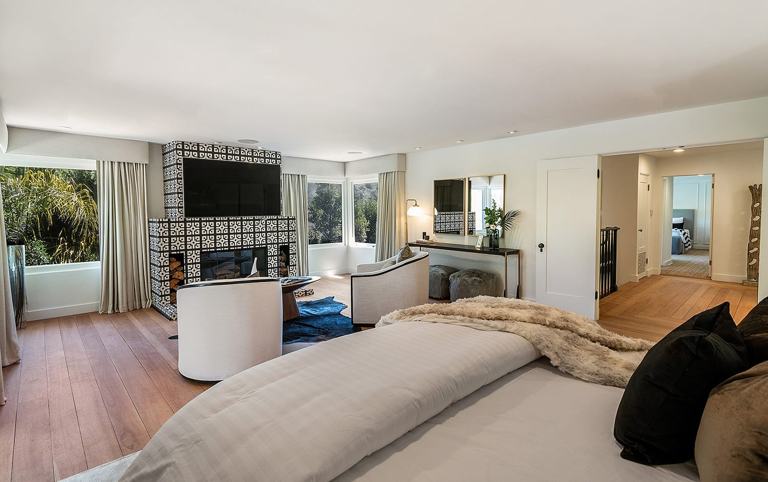 Modern bedroom with a cozy bed, elegant armchairs by a tiled fireplace, large windows, and a scenic view.