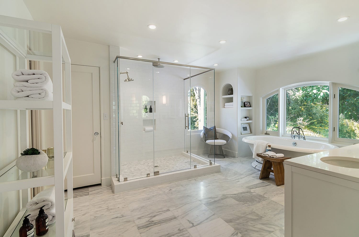 Spacious bathroom with glass shower, marble floors, large window, and shelving with towels.