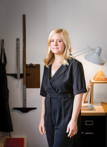 Person standing in an office wearing a black outfit, with a lamp and desk in the background.