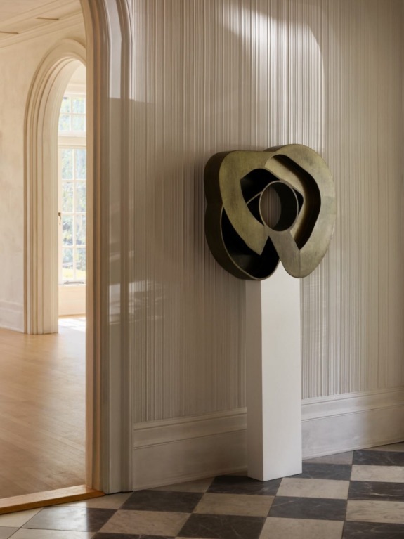 Abstract sculpture on a tall white pedestal in a sunlit room with checkered floor and open doorway.