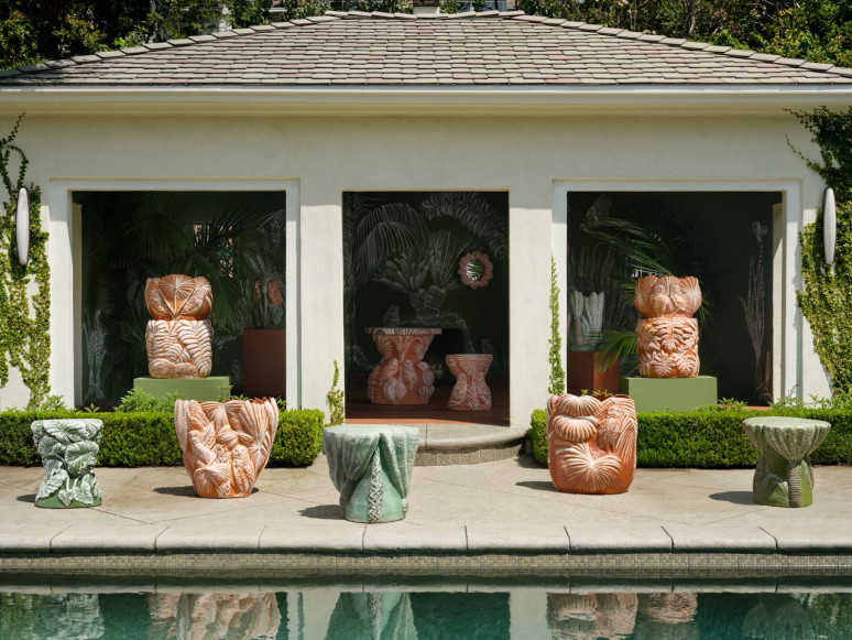 Colorful abstract sculptures displayed by a poolside patio against a lush backdrop of green foliage and a white building.