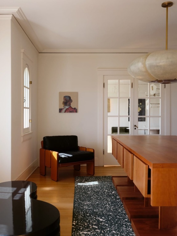 Modern dining area with a large wooden table, black chair, elegant hanging light fixture, and a small wall art piece.