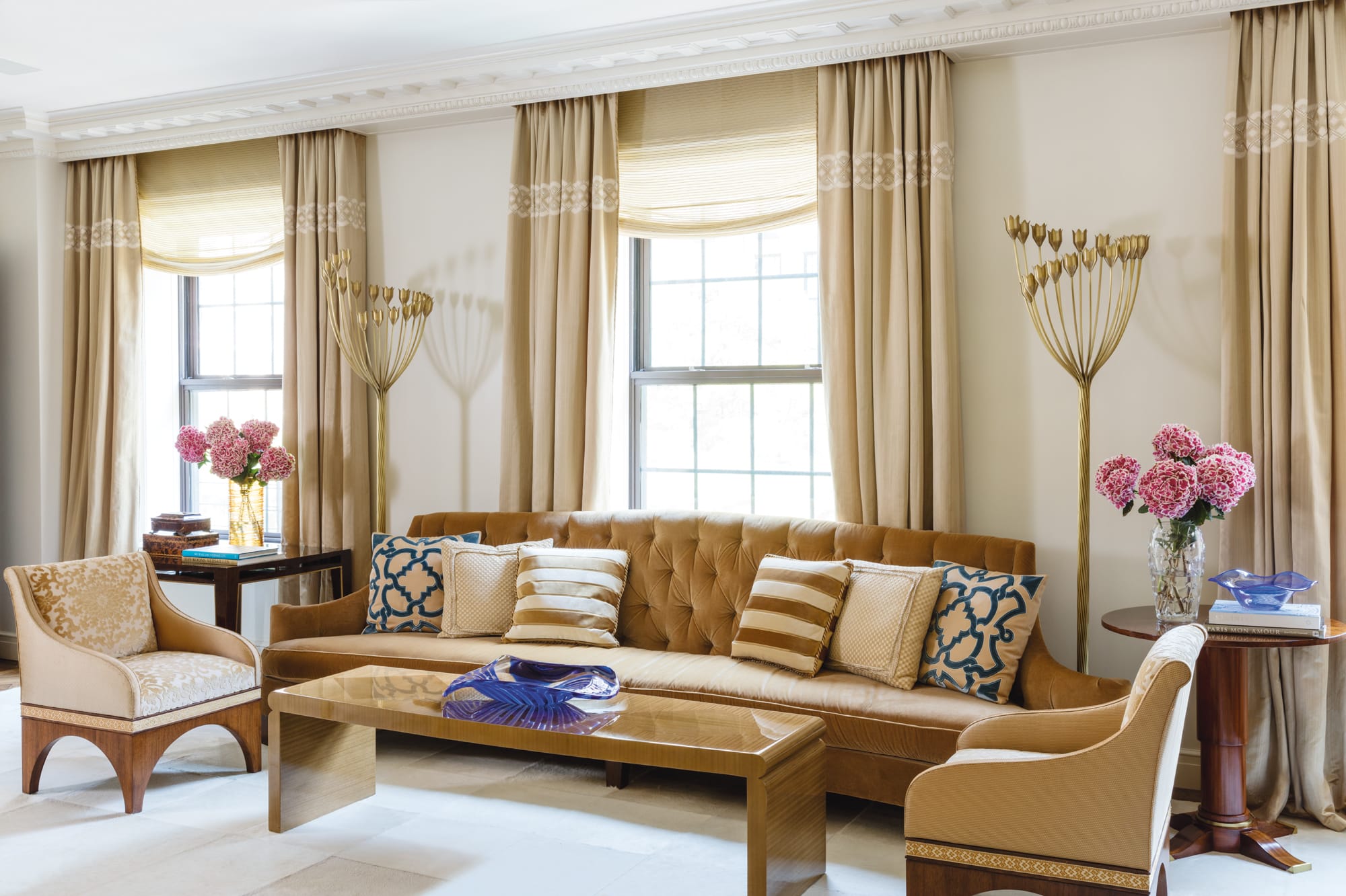 Elegant living room with beige curtains, modern sofa, patterned pillows, and pink flower arrangements on tables.