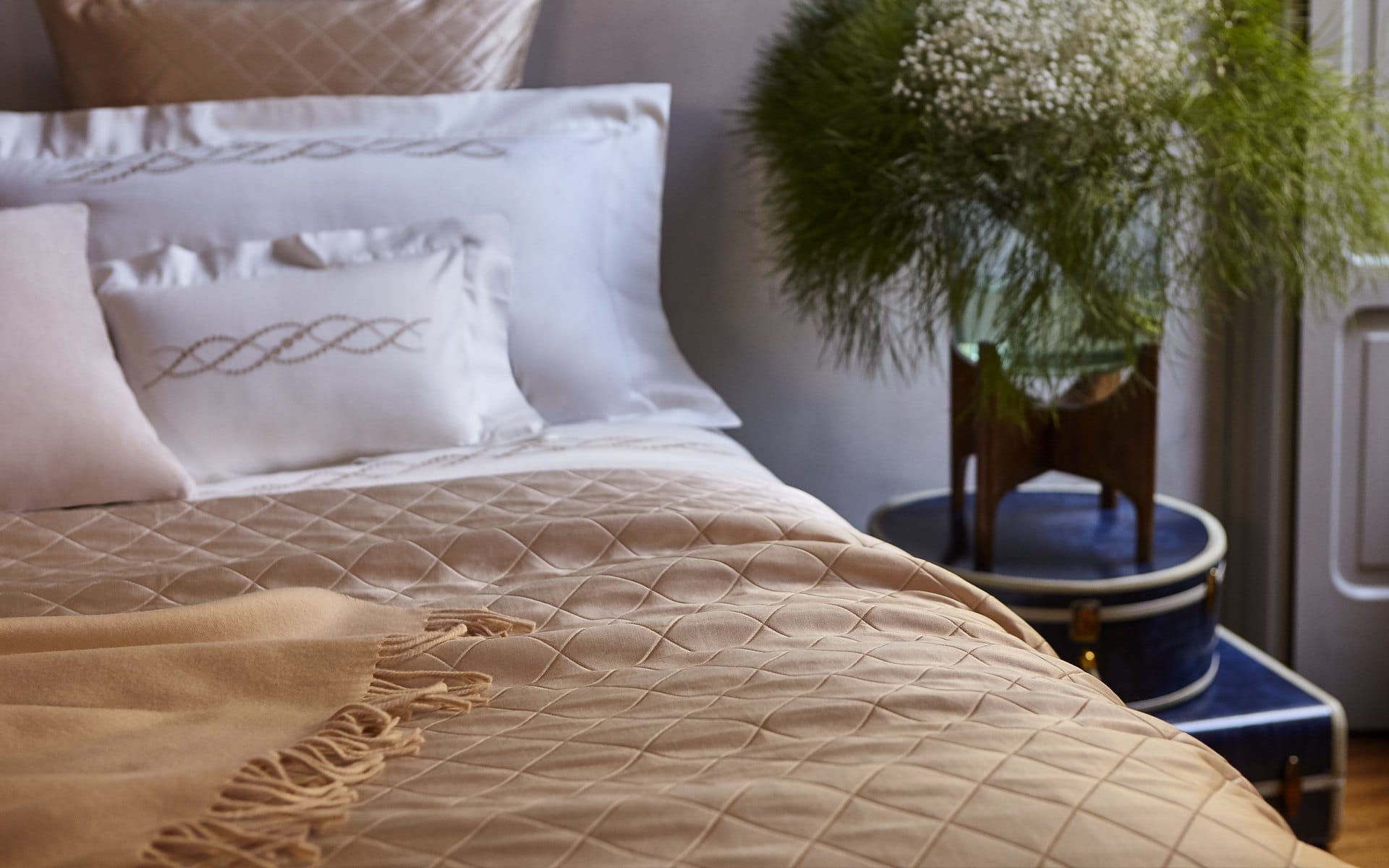 Cozy bed with elegant beige bedding and pillows, next to a small table with a lush green plant arrangement.
